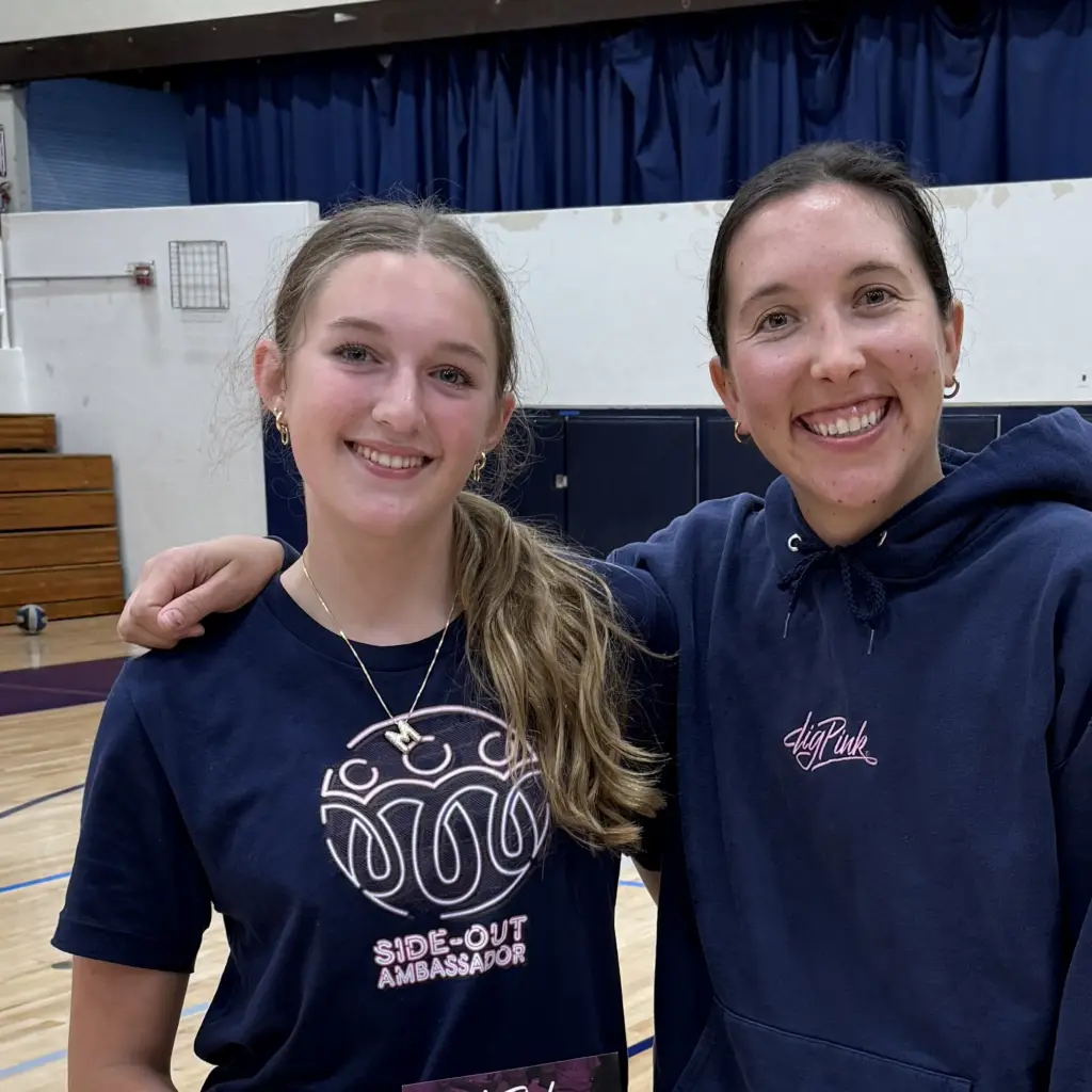 Two young women smile for a selfie in a gym, wearing matching navy shirts that read 'Side-Out Ambassador' with one arm around the other.